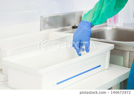 Gloved hand in a sterile protective gown cleans a surgical instrument in a white plastic sterilization tray to maintain safety protocols within a modern medical laboratory environment 140672972