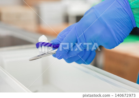 Gloved hand cleans a surgical instrument with a small scrubbing brush in a sterile hospital setting, representing the essential maintenance of medical equipment for patient safety Gloved hand cleans a surgical instrument with a small scrubbing brush in a sterile hospital setting, representing the essential maintenance of medical equipment for patient safety 140672973