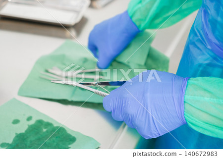 Medical professional wearing blue surgical gloves handles sterile metal instruments on a green drape, depicting focused preparation for a clinical procedure in a hospital setting Medical professional wearing blue surgical gloves handles sterile metal instruments on a green drape, depicting focused preparation for a clinical procedure in a hospital setting 140672983