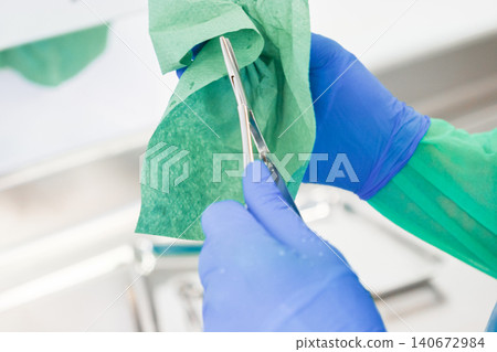 A medical professional wearing blue latex gloves wipes a stainless steel surgical tool with a sterile green paper towel in a hospital to ensure safety during daily clinical tasks A medical professional wearing blue latex gloves wipes a stainless steel surgical tool with a sterile green paper towel in a hospital to ensure safety during daily clinical tasks 140672984