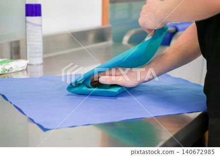 A person folds a sterile blue medical drape on a metallic hospital counter to prepare for a surgical procedure, emphasizing professional cleanliness, clinical safety, and care A person folds a sterile blue medical drape on a metallic hospital counter to prepare for a surgical procedure, emphasizing professional cleanliness, clinical safety, and care 140672985
