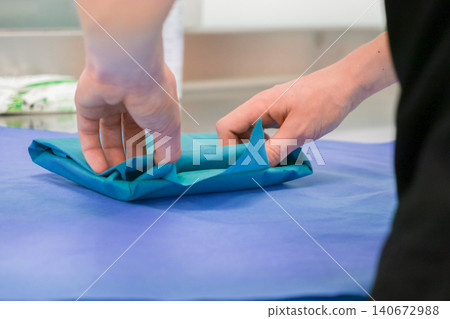 Medical staff member folding blue surgical sterile drape on table for operation prep representing professional hospital hygiene protocols and safety standards in clinical environment Medical staff member folding blue surgical sterile drape on table for operation prep representing professional hospital hygiene protocols and safety standards in clinical environment 140672988