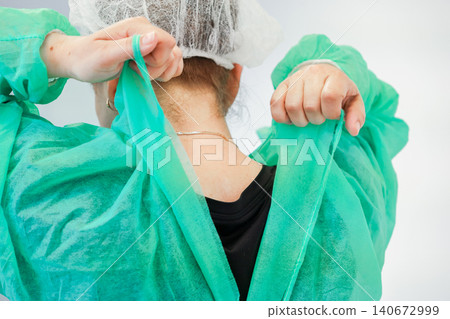A medical professional puts on a green surgical gown and disposable hair cap to demonstrate proper safety protocols for infection control within a clinical laboratory environment 140672999