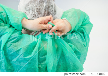 A medical professional ties the back of a green protective surgical gown and wears a bouffant cap in a clinical hospital setting to maintain essential hygiene and safety protocols A medical professional ties the back of a green protective surgical gown and wears a bouffant cap in a clinical hospital setting to maintain essential hygiene and safety protocols 140673002