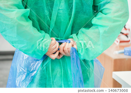 A medical professional carefully ties a blue protective apron over a green gown to demonstrate essential clinical preparation and strict patient safety protocols in a hospital A medical professional carefully ties a blue protective apron over a green gown to demonstrate essential clinical preparation and strict patient safety protocols in a hospital 140673008
