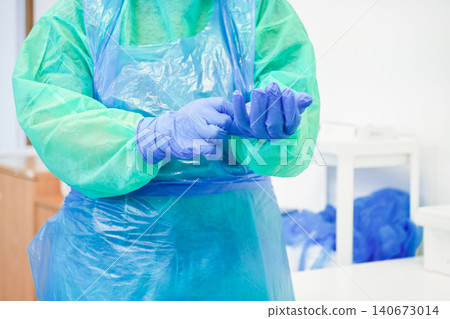 A medical worker puts on blue nitrile protective gloves while wearing a green gown and plastic apron in a clinic to signify rigorous hygiene standards for patient safety and health A medical worker puts on blue nitrile protective gloves while wearing a green gown and plastic apron in a clinic to signify rigorous hygiene standards for patient safety and health 140673014