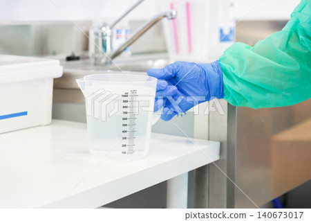 Gloved hand holding a clear plastic measuring jug with liquid in a professional laboratory, reflecting sterile scientific procedures and accurate chemical preparation in a research site Gloved hand holding a clear plastic measuring jug with liquid in a professional laboratory, reflecting sterile scientific procedures and accurate chemical preparation in a research site 140673017