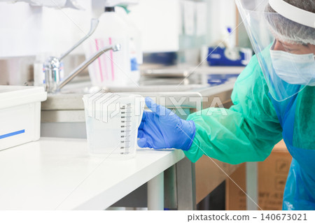 Medical professional wearing protective gear holds a plastic beaker in a sterile laboratory setting, demonstrating clinical precision during a controlled scientific research procedure 140673021