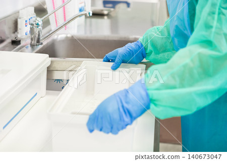 A medical professional in protective gear handles a sterile plastic container near a stainless steel sink in a clinical lab to ensure high hygiene standards during equipment prep A medical professional in protective gear handles a sterile plastic container near a stainless steel sink in a clinical lab to ensure high hygiene standards during equipment prep 140673047