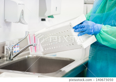 A gloved worker sanitizing a plastic basket at a sink in a medical facility to emphasize the strict hygiene protocols and cleaning procedures required for a sterile environment A gloved worker sanitizing a plastic basket at a sink in a medical facility to emphasize the strict hygiene protocols and cleaning procedures required for a sterile environment 140673050