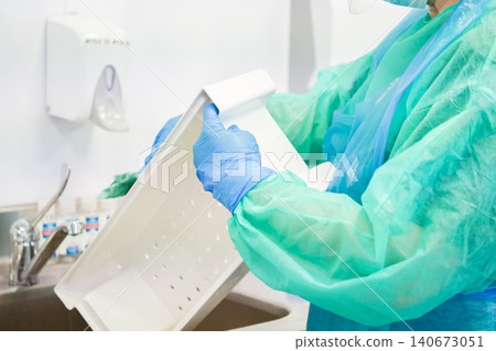 A dedicated healthcare worker in protective gear washes a white medical tray in a clinic sink to emphasize essential sanitation procedures and hygiene maintenance for patient safety A dedicated healthcare worker in protective gear washes a white medical tray in a clinic sink to emphasize essential sanitation procedures and hygiene maintenance for patient safety 140673051