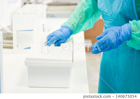 Medical worker in protective apron and blue gloves opens a sterile white plastic container at a clinic table, ensuring hygiene and safety standards in a professional lab environment 140673054