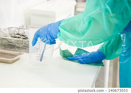 A medical staff member wearing a surgical gown and blue gloves cleans a white laboratory surface to maintain strict hygiene standards and ensure a sterile clinical environment A medical staff member wearing a surgical gown and blue gloves cleans a white laboratory surface to maintain strict hygiene standards and ensure a sterile clinical environment 140673055