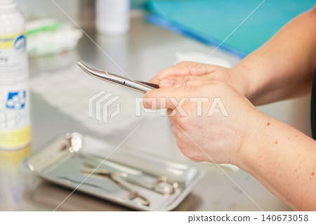 A dental assistant holds stainless steel forceps over a metal tray in a clinic room, illustrating precise preparation for an oral surgical procedure within a medical setting 140673058