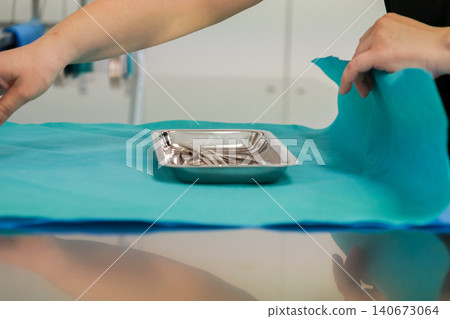 A medical professional wraps sterile steel surgical instruments in a green cloth for hospital procedure setup in a clinical setting to ensure safety for a patient operation A medical professional wraps sterile steel surgical instruments in a green cloth for hospital procedure setup in a clinical setting to ensure safety for a patient operation 140673064