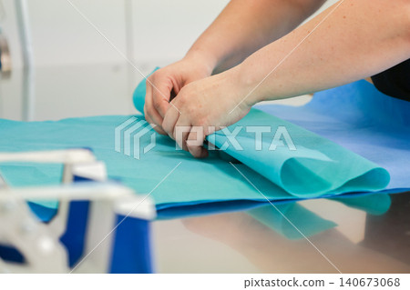 Medical professional hands folding sterile blue surgical paper on a metallic table, representing hygiene, clinical preparation, equipment maintenance, and safety in hospitals Medical professional hands folding sterile blue surgical paper on a metallic table, representing hygiene, clinical preparation, equipment maintenance, and safety in hospitals 140673068