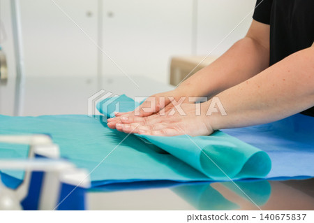 Medical professional folds sterile blue surgical drape on a metal table in a clinical setting to ensure patient safety and maintain an aseptic environment for surgery preparation Medical professional folds sterile blue surgical drape on a metal table in a clinical setting to ensure patient safety and maintain an aseptic environment for surgery preparation 140675837