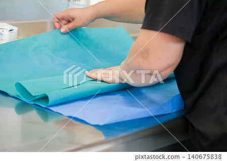 Medical professional hands preparing sterile blue disposable paper sheets on stainless table, ensuring clean equipment arrangement for surgical procedures in a modern hospital Medical professional hands preparing sterile blue disposable paper sheets on stainless table, ensuring clean equipment arrangement for surgical procedures in a modern hospital 140675838