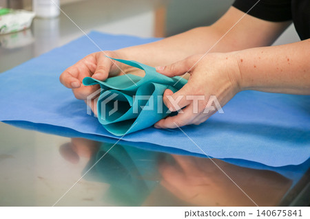 A medical professional carefully folds a sterile blue surgical drape on a metal table for an upcoming operation to ensure patient safety and maintain a clean hospital clinic room A medical professional carefully folds a sterile blue surgical drape on a metal table for an upcoming operation to ensure patient safety and maintain a clean hospital clinic room 140675841