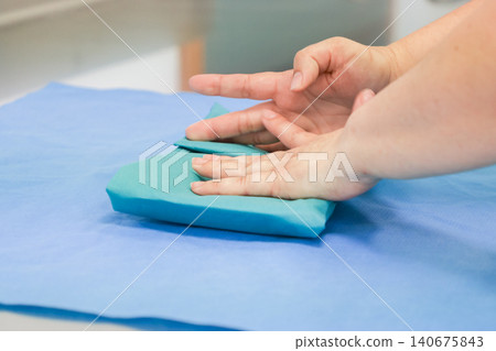 A medical professional folds a sterile blue surgical drape on a table, maintaining strict hygiene protocols to ensure safety and precision within a clinical hospital operating room A medical professional folds a sterile blue surgical drape on a table, maintaining strict hygiene protocols to ensure safety and precision within a clinical hospital operating room 140675843