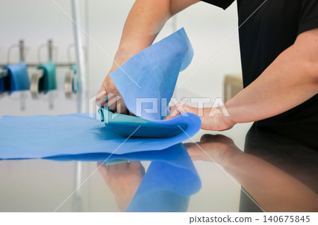 Medical professional hands wrapping sterile surgical instruments in blue nonwoven fabric at a stainless steel table to ensure clinical hygiene and safety in a hospital setting Medical professional hands wrapping sterile surgical instruments in blue nonwoven fabric at a stainless steel table to ensure clinical hygiene and safety in a hospital setting 140675845