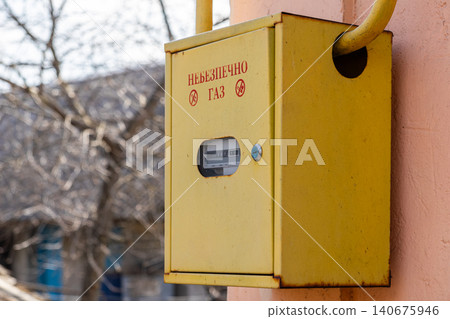 Yellow metal gas meter box attached to a residential wall with a warning sign, representing essential utility infrastructure safety and hazard prevention in a rural living area Yellow metal gas meter box attached to a residential wall with a warning sign, representing essential utility infrastructure safety and hazard prevention in a rural living area 140675946