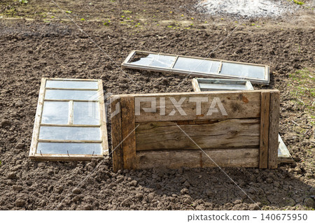 Old wooden window frames and panels resting on dark plowed soil in a garden, creating a rustic textured scene that highlights abandoned materials and preparation for spring planting Old wooden window frames and panels resting on dark plowed soil in a garden, creating a rustic textured scene that highlights abandoned materials and preparation for spring planting 140675950