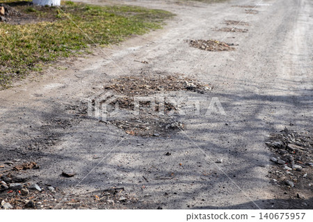 A broken asphalt road displays multiple deep potholes and cracks across the surface, highlighting poor urban infrastructure, neglected maintenance, and hazardous suburban travel A broken asphalt road displays multiple deep potholes and cracks across the surface, highlighting poor urban infrastructure, neglected maintenance, and hazardous suburban travel 140675957