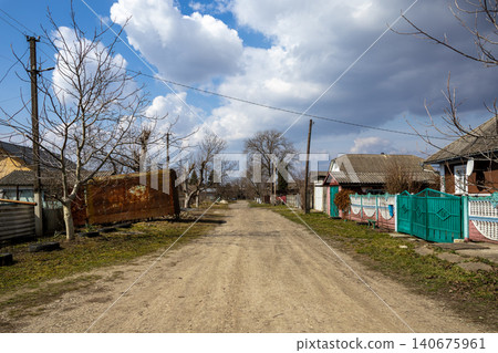Unpaved dirt road leading through a quiet rural village with traditional small houses and bare trees under a bright blue sky, representing simple countryside life and remote areas Unpaved dirt road leading through a quiet rural village with traditional small houses and bare trees under a bright blue sky, representing simple countryside life and remote areas 140675961