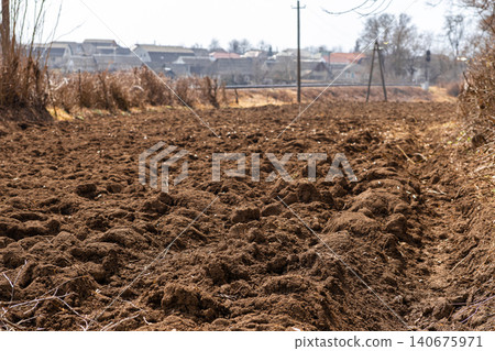 Freshly plowed soil in a rural field with a distant village in the background, conveying spring agricultural preparation and the essence of traditional farming life in the countryside Freshly plowed soil in a rural field with a distant village in the background, conveying spring agricultural preparation and the essence of traditional farming life in the countryside 140675971