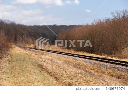 Curving train tracks stretch through a scenic rural autumn landscape under a bright blue sky, highlighting transport infrastructure amidst quiet woods and dry grassy terrain now Curving train tracks stretch through a scenic rural autumn landscape under a bright blue sky, highlighting transport infrastructure amidst quiet woods and dry grassy terrain now 140675974