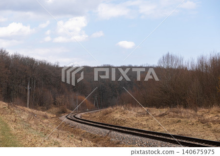 Curving railroad tracks leading into a dense autumn forest under a cloudy blue sky, capturing the quiet solitude and steady progress of transportation through a natural landscape Curving railroad tracks leading into a dense autumn forest under a cloudy blue sky, capturing the quiet solitude and steady progress of transportation through a natural landscape 140675975