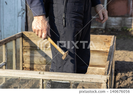 A senior man holds a vintage hammer over a wooden garden frame, representing manual labor, small home improvement projects, and active outdoor property maintenance during the dusk 140676056