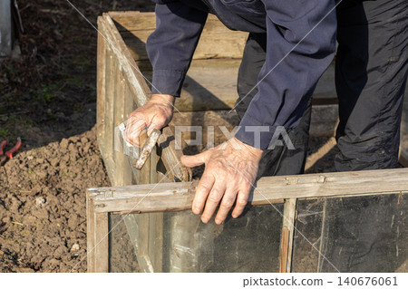 Person using a metal hammer to assemble a wooden frame greenhouse in a sunny garden, capturing the process of DIY backyard improvement for sustainable plant growth and cultivation Person using a metal hammer to assemble a wooden frame greenhouse in a sunny garden, capturing the process of DIY backyard improvement for sustainable plant growth and cultivation 140676061