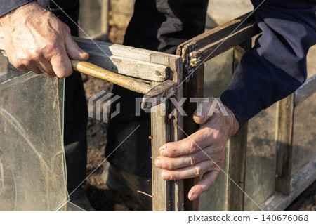 A senior man hammers a metal nail into the weathered wooden frame of a vintage garden greenhouse, demonstrating precision in outdoor construction and manual repair work today A senior man hammers a metal nail into the weathered wooden frame of a vintage garden greenhouse, demonstrating precision in outdoor construction and manual repair work today 140676068
