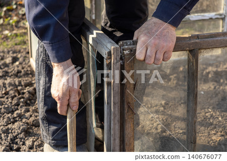 A person carefully assembles a wooden garden cold frame with glass panels over soil to protect plants, creating an organized outdoor space for spring planting and home gardening A person carefully assembles a wooden garden cold frame with glass panels over soil to protect plants, creating an organized outdoor space for spring planting and home gardening 140676077