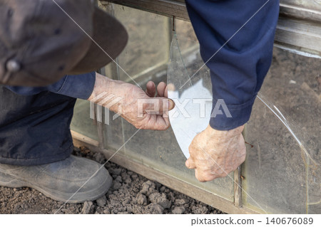 Senior man repairing a broken glass pane on a wooden garden greenhouse frame with careful hands, representing manual labor and home maintenance work in an outdoor spring setting Senior man repairing a broken glass pane on a wooden garden greenhouse frame with careful hands, representing manual labor and home maintenance work in an outdoor spring setting 140676089