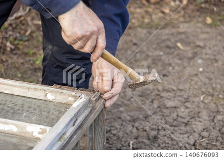 A person hammers a small metal nail into a weathered wooden frame outdoors to represent DIY home improvement, manual labor, craftsmanship, and the dedicated restoration of furniture A person hammers a small metal nail into a weathered wooden frame outdoors to represent DIY home improvement, manual labor, craftsmanship, and the dedicated restoration of furniture 140676093