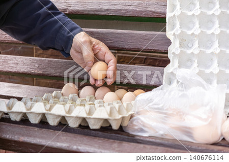 A person places a fresh organic brown egg into a paper carton on a wooden bench to capture the simple essence of farm to table food preparation and sustainable rural lifestyles A person places a fresh organic brown egg into a paper carton on a wooden bench to capture the simple essence of farm to table food preparation and sustainable rural lifestyles 140676114