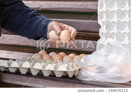 A person holds fresh brown chicken eggs near a cardboard carton on a wooden bench, representing organic farming, rural food production, and a sustainable local healthy diet A person holds fresh brown chicken eggs near a cardboard carton on a wooden bench, representing organic farming, rural food production, and a sustainable local healthy diet 140676115