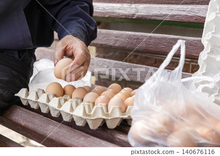 Older person holding a fresh brown chicken egg over a cardboard carton on a wooden bench, representing organic farm produce, food shopping, and sustainable healthy living at home 140676116