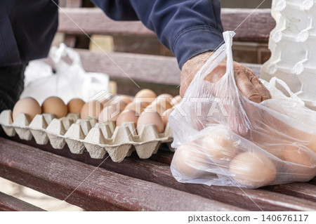 Person placing fresh brown eggs into a plastic bag from a paper carton on a wooden bench, conveying local farm shopping and healthy organic food choices in a simple daily lifestyle 140676117