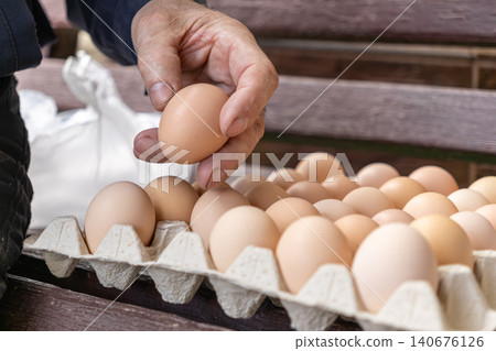 A person hand picks a fresh brown egg from a cardboard carton to represent organic farm produce selection, sustainable food sourcing, and rustic natural ingredients for cooking A person hand picks a fresh brown egg from a cardboard carton to represent organic farm produce selection, sustainable food sourcing, and rustic natural ingredients for cooking 140676126