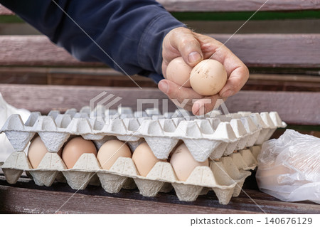 A hand holds raw organic chicken eggs above a cardboard carton tray on a rustic wooden bench to demonstrate local farming harvest, sustainability, and fresh agricultural food A hand holds raw organic chicken eggs above a cardboard carton tray on a rustic wooden bench to demonstrate local farming harvest, sustainability, and fresh agricultural food 140676129