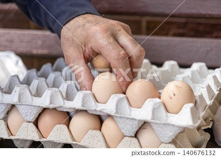 Older person hand picking raw brown chicken egg from paper carton crate, symbolizing fresh organic farm produce, sustainable food sourcing, healthy lifestyle, and culinary activity Older person hand picking raw brown chicken egg from paper carton crate, symbolizing fresh organic farm produce, sustainable food sourcing, healthy lifestyle, and culinary activity 140676132
