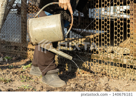 A person holds a vintage metal watering can to pour water onto dry soil in a rural garden, capturing the essence of manual labor and traditional sustainable backyard farming habits A person holds a vintage metal watering can to pour water onto dry soil in a rural garden, capturing the essence of manual labor and traditional sustainable backyard farming habits 140676181