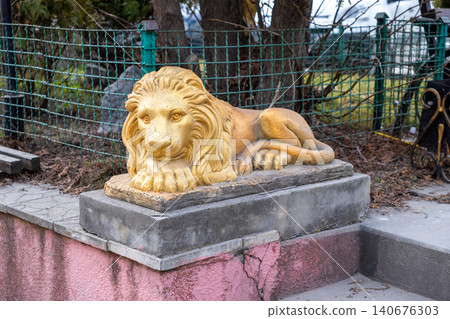 A golden lion statue rests on a concrete pedestal in a garden, providing an elegant decorative element that adds artistic flair to the landscape design of a residential property yard A golden lion statue rests on a concrete pedestal in a garden, providing an elegant decorative element that adds artistic flair to the landscape design of a residential property yard 140676303