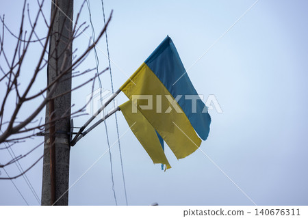 A weathered flag of Ukraine hangs from a tall utility pole against a soft gray sky, symbolizing resilience, national pride, and strength during challenging geopolitical times A weathered flag of Ukraine hangs from a tall utility pole against a soft gray sky, symbolizing resilience, national pride, and strength during challenging geopolitical times 140676311