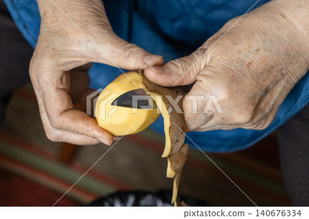 Elderly hands peeling a fresh raw potato with a small kitchen knife, emphasizing traditional home food preparation skills and domestic culinary tasks in a quiet household setting Elderly hands peeling a fresh raw potato with a small kitchen knife, emphasizing traditional home food preparation skills and domestic culinary tasks in a quiet household setting 140676334
