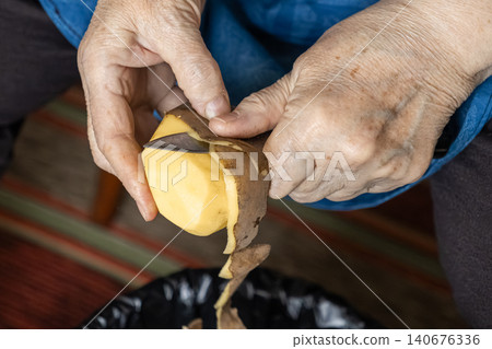 A senior person peels a raw potato with a small steel knife, representing traditional home cooking preparation and domestic life in a serene atmosphere of culinary skill and care A senior person peels a raw potato with a small steel knife, representing traditional home cooking preparation and domestic life in a serene atmosphere of culinary skill and care 140676336